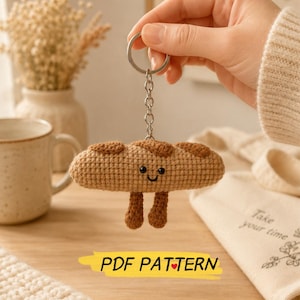 May include: A crocheted baguette keyring featuring a light brown textured body with darker brown accents, two small dangling legs, and a simple embroidered face with black eyes and a smile. The text PDF PATTERN is printed below.