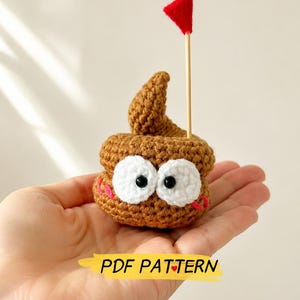 May include: A brown crocheted figure with a swirled top, two large white eyes with black pupils, and pink stitched cheeks. A thin wooden stick with a small red triangular flag is inserted into the top. The text PDF PATTERN is visible.