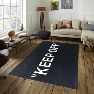 May include: A black rectangular rug with the words "KEEP OFF" in large white capital letters. The rug is placed on a hardwood floor in a living room setting, with a sofa, side table, and window visible in the background.