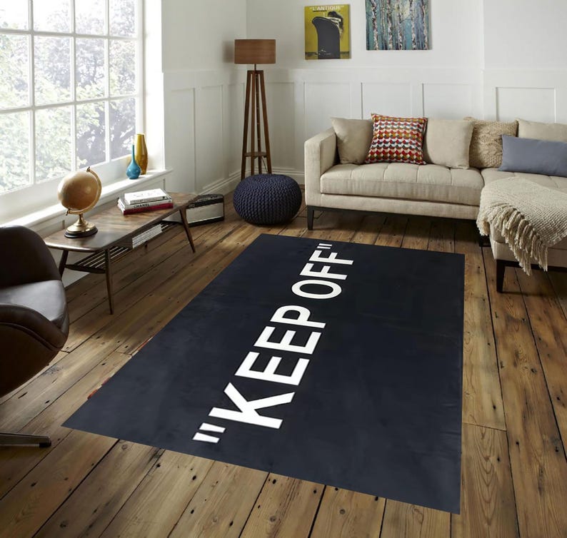 May include: A black rectangular rug with the words "KEEP OFF" in large white capital letters. The rug is placed on a hardwood floor in a living room setting, with a sofa, side table, and window visible in the background.