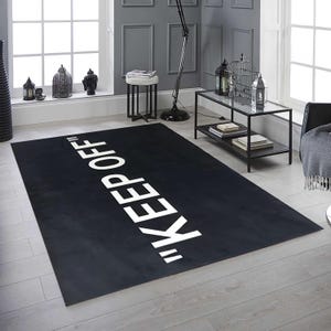 May include: A rectangular black rug with the words "KEEP OFF" in large white capital letters. The rug is placed on a light-colored wooden floor in a room with gray walls and a window.