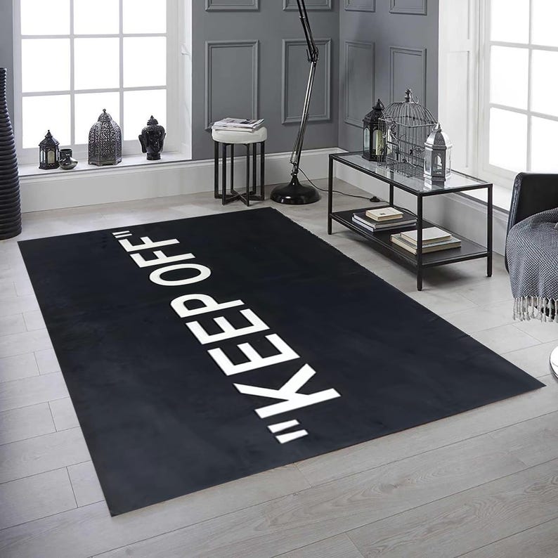 May include: A rectangular black rug with the words "KEEP OFF" in large white capital letters. The rug is placed on a light-colored wooden floor in a room with gray walls and a window.