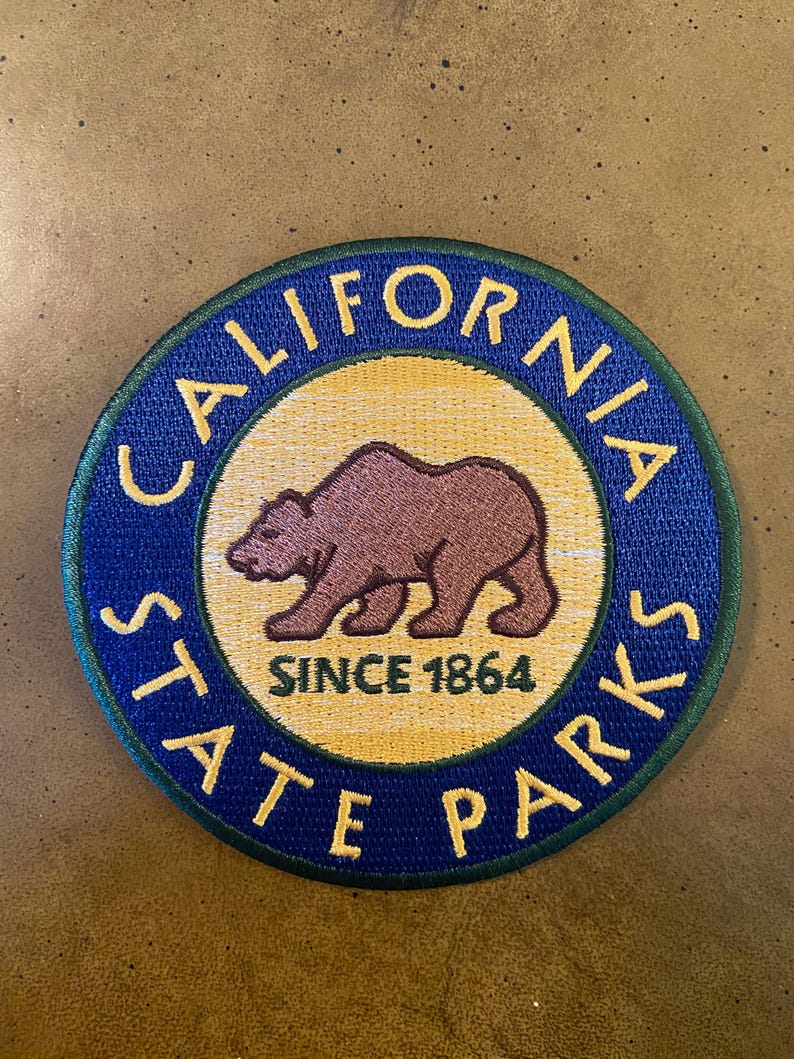 May include: A blue and yellow round patch with the text "California State Parks" and a brown bear silhouette. The text "Since 1864" is inside the circle.