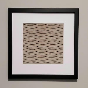 May include: A framed artwork featuring a textured, wave-like pattern in shades of beige and brown. The artwork is set within a white mat and a black frame, creating a modern and minimalist aesthetic. The piece is suitable for home decor.