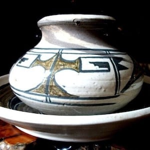 May include: A white ceramic pot with a brown, black, and white geometric design. The pot is sitting on a white ceramic plate.
