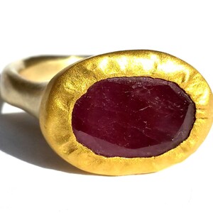 May include: A gold ring featuring a large, oval ruby gemstone set in a textured gold bezel. The ring is crafted from 14k gold and has a hammered finish.