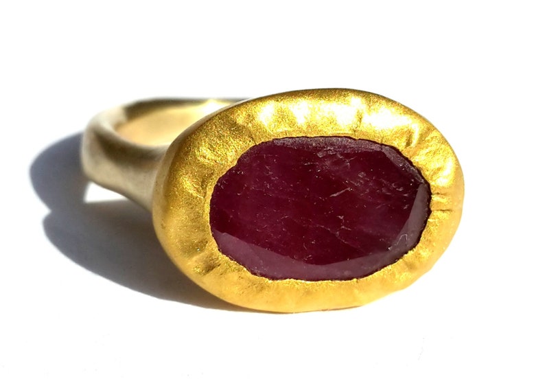 May include: A gold ring featuring a large, oval ruby gemstone set in a textured gold bezel. The ring is crafted from 14k gold and has a hammered finish.