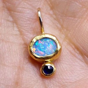 Op de afbeelding: Een gouden hanger met een ovale opaal met blauwe en iriserende tinten, boven een kleine, ronde, donkerblauwe edelsteen. De hanger heeft een lus aan de bovenkant voor een ketting.