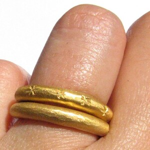 May include: Two gold rings stacked on a finger. The top ring has a textured surface with small star-shaped designs. The bottom ring is smooth.