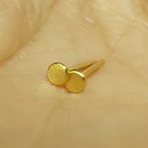 Puede incluir: Un par de peque&ntilde;os pendientes de bot&oacute;n redondos de color dorado. Los pendientes tienen una superficie texturizada y se muestran sobre un fondo marr&oacute;n claro.