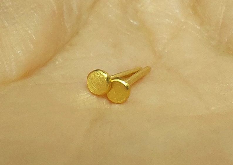 Puede incluir: Un par de peque&ntilde;os pendientes de bot&oacute;n redondos de color dorado. Los pendientes tienen una superficie texturizada y se muestran sobre un fondo marr&oacute;n claro.