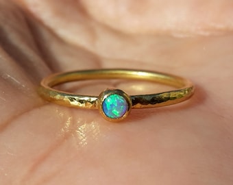 May include: A delicate gold ring with a hammered band and a small, round opal gemstone in the center. The opal has a blue-green iridescent color.