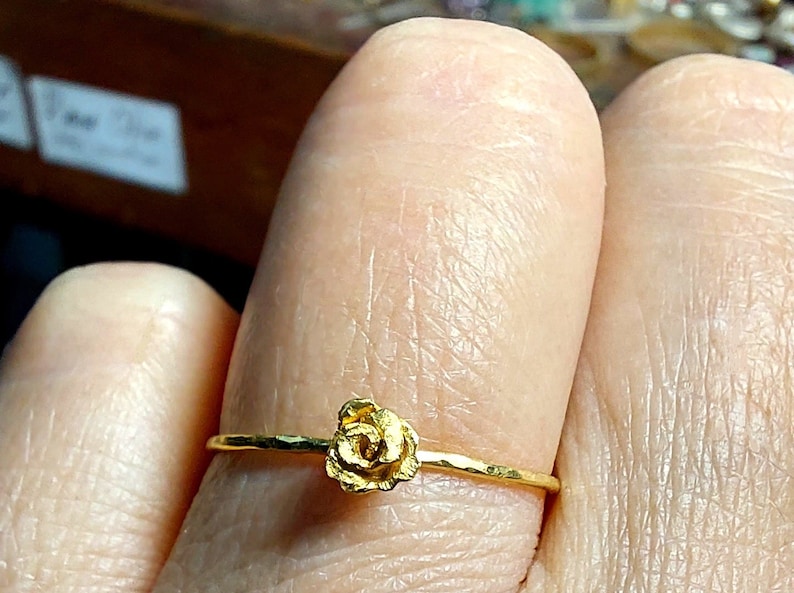 May include: A delicate gold ring with a small rose-shaped design. The ring is worn on a finger.