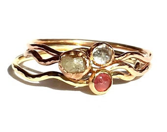 May include: Three gold rings with different coloured gemstones. The rings are stacked on top of each other and have a wavy design. The gemstones are a light pink, a clear white, and a light yellow.
