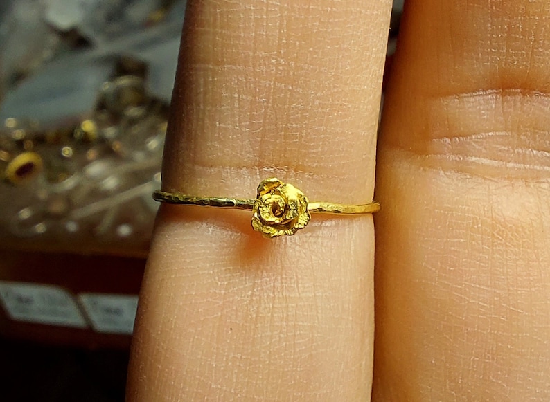 May include: A delicate gold ring with a small rose design. The ring is worn on a finger.