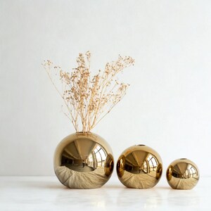 May include: Three gold spherical vases of varying sizes. The largest vase holds dried, light brown stems. The vases have a reflective, metallic finish, and sit on a white surface against a white background. The vases are decorative.