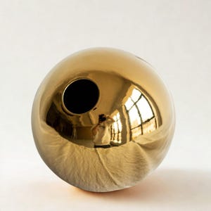 May include: A spherical, gold-colored vase with a glossy finish. The vase has a single, circular opening and reflects the surrounding environment. The surface is smooth and reflective, creating a mirror-like effect.