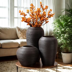 Può includere: Tre vasi marrone scuro strutturati di diverse dimensioni sono esposti su un tavolo rotondo in legno. Il vaso più grande contiene rami arancioni. I vasi hanno un design moderno, adatti per l'arredamento della casa.