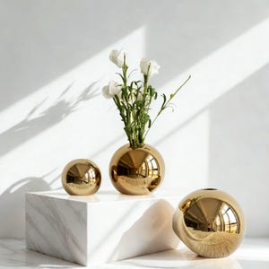 May include: Three gold spherical vases of varying sizes. The largest vase holds white flowers. The vases are displayed on a white marble block against a white background. The vases have a reflective, metallic finish.