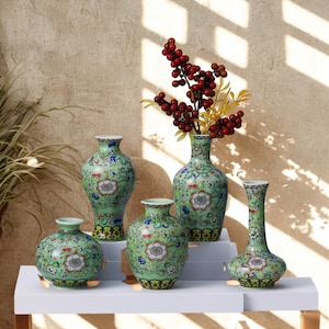 May include: Five green ceramic vases with floral designs. The vases are of different shapes and sizes, including round and elongated forms. One vase contains red berries and yellow sprigs. The vases are displayed on a white surface.