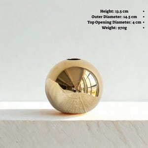 May include: A spherical, gold-colored vase with a reflective surface. The vase has a height of 5.3 inches, an outer diameter of 5.7 inches, and a top opening diameter of 1.6 inches. It weighs 34.2 ounces. The vase is sitting on a white surface.