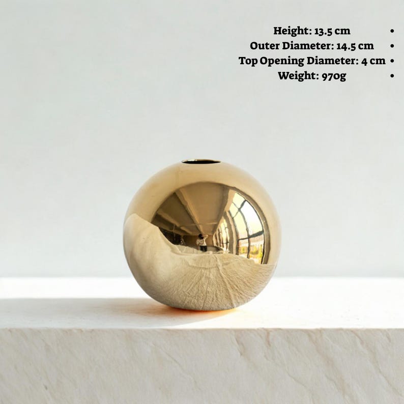 May include: A spherical, gold-colored vase with a reflective surface. The vase has a height of 5.3 inches, an outer diameter of 5.7 inches, and a top opening diameter of 1.6 inches. It weighs 34.2 ounces. The vase is sitting on a white surface.