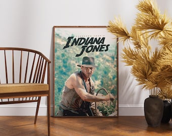 May include: A framed poster featuring the movie title "Indiana Jones" with Harrison Ford. The poster has a brown frame and is displayed against a white wall. A wooden chair and decorative plants are in the foreground.