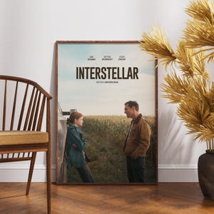 May include: Framed movie poster for "Interstellar" with a brown wooden frame. The poster features two people standing in a field, with the title "Interstellar" in large letters. A wooden chair and decorative vase with dried plants are in the foreground.