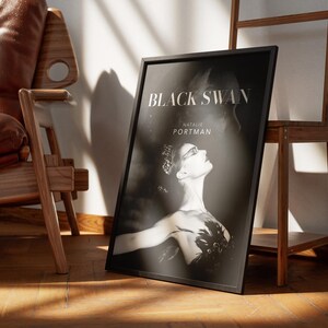 May include: Black and white framed poster for the movie "Black Swan" featuring Natalie Portman. The poster is propped against a wooden easel and a wall, with a leather chair in the background. The poster has a black frame.