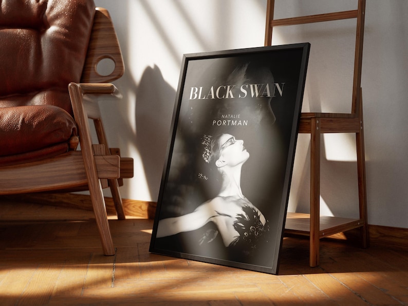 May include: Black and white framed poster for the movie "Black Swan" featuring Natalie Portman. The poster is propped against a wooden easel and a wall, with a leather chair in the background. The poster has a black frame.