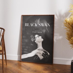 May include: A framed poster for the movie "Black Swan" featuring a black and white image of Natalie Portman. The poster has a dark wooden frame and is leaning against a white wall. A wooden chair and decorative dried plants are in the foreground.