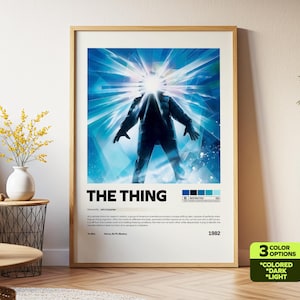 May include: Framed movie poster for "The Thing" featuring a figure with a bright light emanating from its head, set against a blue and white background. The title is in bold white letters. The poster is in a wooden frame.