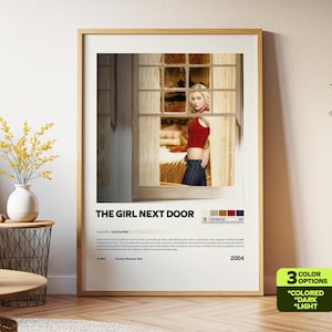 May include: Framed movie poster for "The Girl Next Door" with a blonde woman in a red top and jeans. The poster has a wooden frame and includes the movie title, credits, and a color palette.