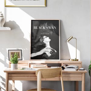 May include: A framed "Black Swan" movie poster featuring Natalie Portman, displayed on a wooden desk. The black and white image is set against a neutral wall, with a small plant, books, and a gold desk lamp. A wooden chair is in front of the desk.