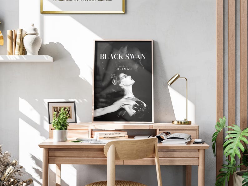 May include: A framed "Black Swan" movie poster featuring Natalie Portman, displayed on a wooden desk. The black and white image is set against a neutral wall, with a small plant, books, and a gold desk lamp. A wooden chair is in front of the desk.