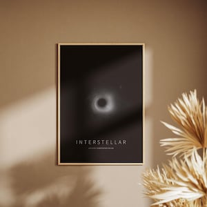 Può includere: Un poster incorniciato con il titolo "INTERSTELLAR" su sfondo nero, con una rappresentazione bianca e grigia di un buco nero. Il poster è in una cornice dorata e appeso a una parete beige.