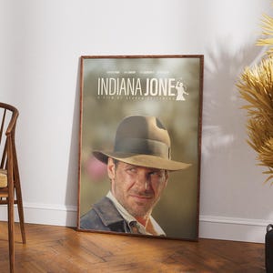 May include: A framed movie poster for "Indiana Jones" featuring a portrait of the main character wearing a fedora. The poster has a brown wooden frame and is leaning against a white wall. The title is displayed at the top.