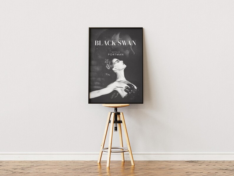 May include: Black and white framed poster featuring the movie title "Black Swan" and the name "Natalie Portman." The image shows a ballerina in a dramatic pose. The poster is displayed on a wooden and metal adjustable stool.