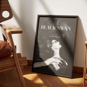 May include: A framed black and white poster for the movie "Black Swan" featuring Natalie Portman. The poster is leaning against a white wall, next to a wooden chair and a small side table. The poster's design is a portrait of a woman with a dark background.