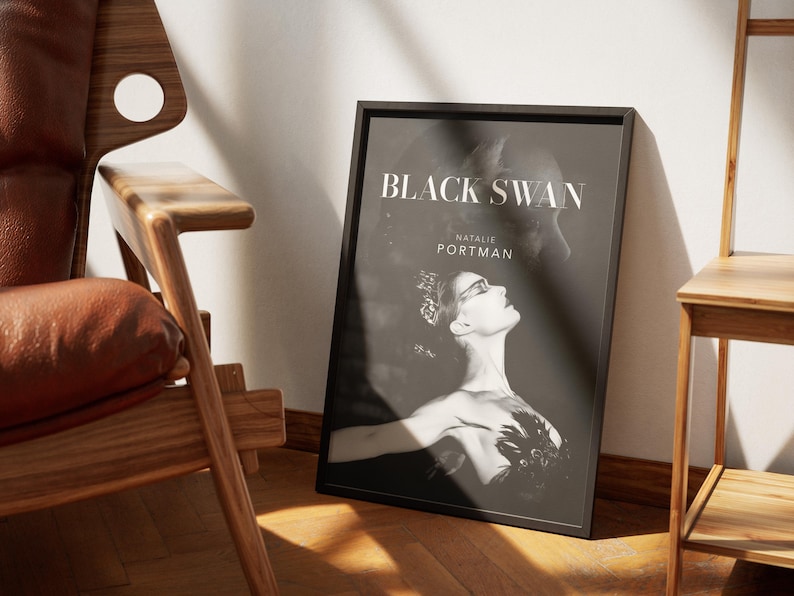 May include: A framed black and white poster for the movie "Black Swan" featuring Natalie Portman. The poster is leaning against a white wall, next to a wooden chair and a small side table. The poster's design is a portrait of a woman with a dark background.