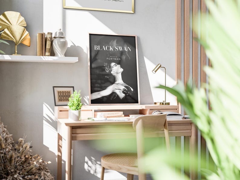 May include: A framed black and white poster of the movie "Black Swan" featuring Natalie Portman, displayed on a wooden desk. The desk has a small lamp, books, and a small plant. A decorative shelf with books and a gold fan is visible.
