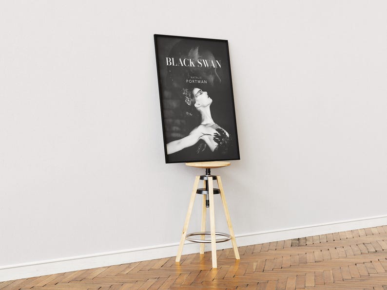 May include: Black and white framed poster of the movie "Black Swan" featuring Natalie Portman. The poster is displayed on a wooden and metal stool against a white wall and a wooden floor.