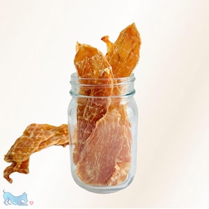 May include: A clear glass mason jar filled with several thin, dried, golden-brown chicken breast strips. One additional dried chicken strip lies horizontally on the surface to the left of the jar. The texture of the dried meat is fibrous and uneven.