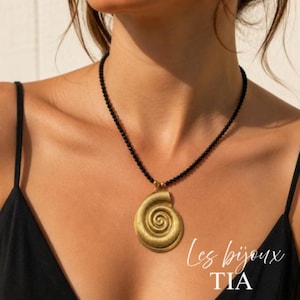 May include: A gold-colored pendant necklace featuring a spiral design, suspended from a black beaded chain. The necklace is worn against a dark top. The text "Les bijoux TIA" is visible in the image.