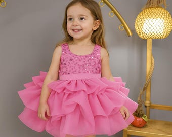 May include: A young girl smiles, wearing a bright pink dress. The sleeveless bodice is embellished with sequins, and the skirt features multiple layers of ruffled fabric. The dress has a matching pink waistband. The girl is standing in front of a gold-colored decorative arch.