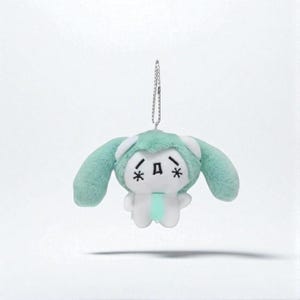 Puede incluir: Un peluche con forma de personaje de dibujos animados con capucha y orejas verde menta, cuerpo blanco y una cadena plateada para colgar. La cara tiene una expresión triste con asteriscos para los ojos y una boca cuadrada.
