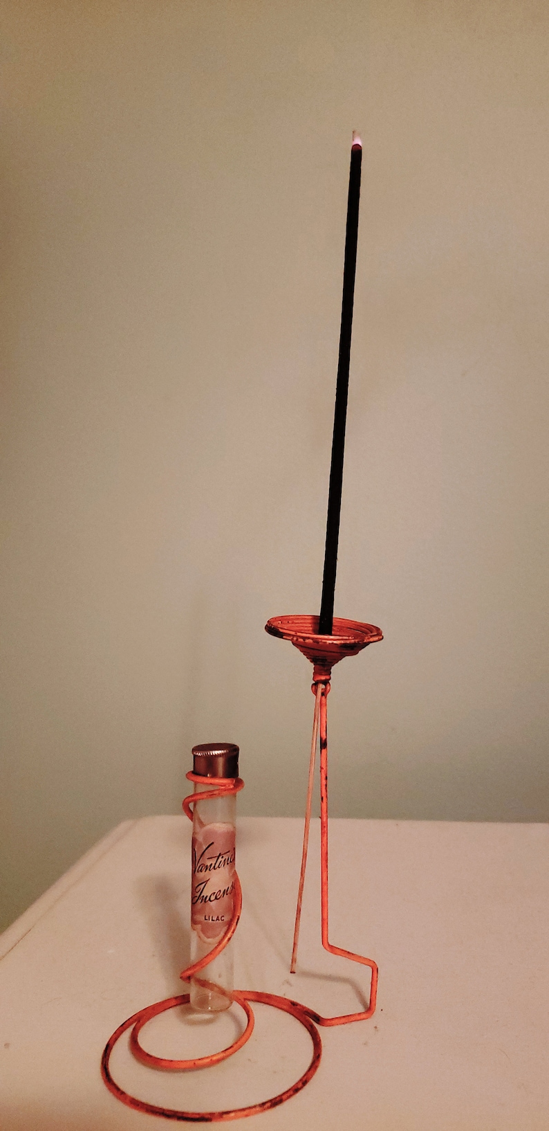 May include: A clear glass bottle with a label that reads "Vantine Incense Lilac" is sitting on a coiled orange wire stand. A black incense stick is burning in a small orange metal holder on a taller orange wire stand.