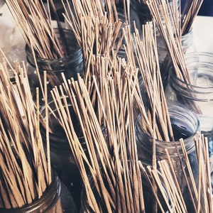 May include: A close-up of several glass jars filled with bundles of thin, brown incense sticks. The jars are arranged in a pyramid shape, with the top jar being the smallest and the bottom jar being the largest. The jars are all clear glass and have a slightly frosted appearance. The incense sticks are all the same length and are bundled together tightly. The sticks are all brown and have a slightly rough texture.