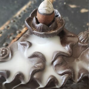 Peut inclure: Un br&ucirc;leur d'encens en c&eacute;ramique brune avec un motif de fum&eacute;e blanche. Le br&ucirc;leur pr&eacute;sente un motif de fleur de lotus avec un porte-encens conique sur le dessus. Le br&ucirc;leur a &eacute;galement deux motifs de poissons sur le fond.