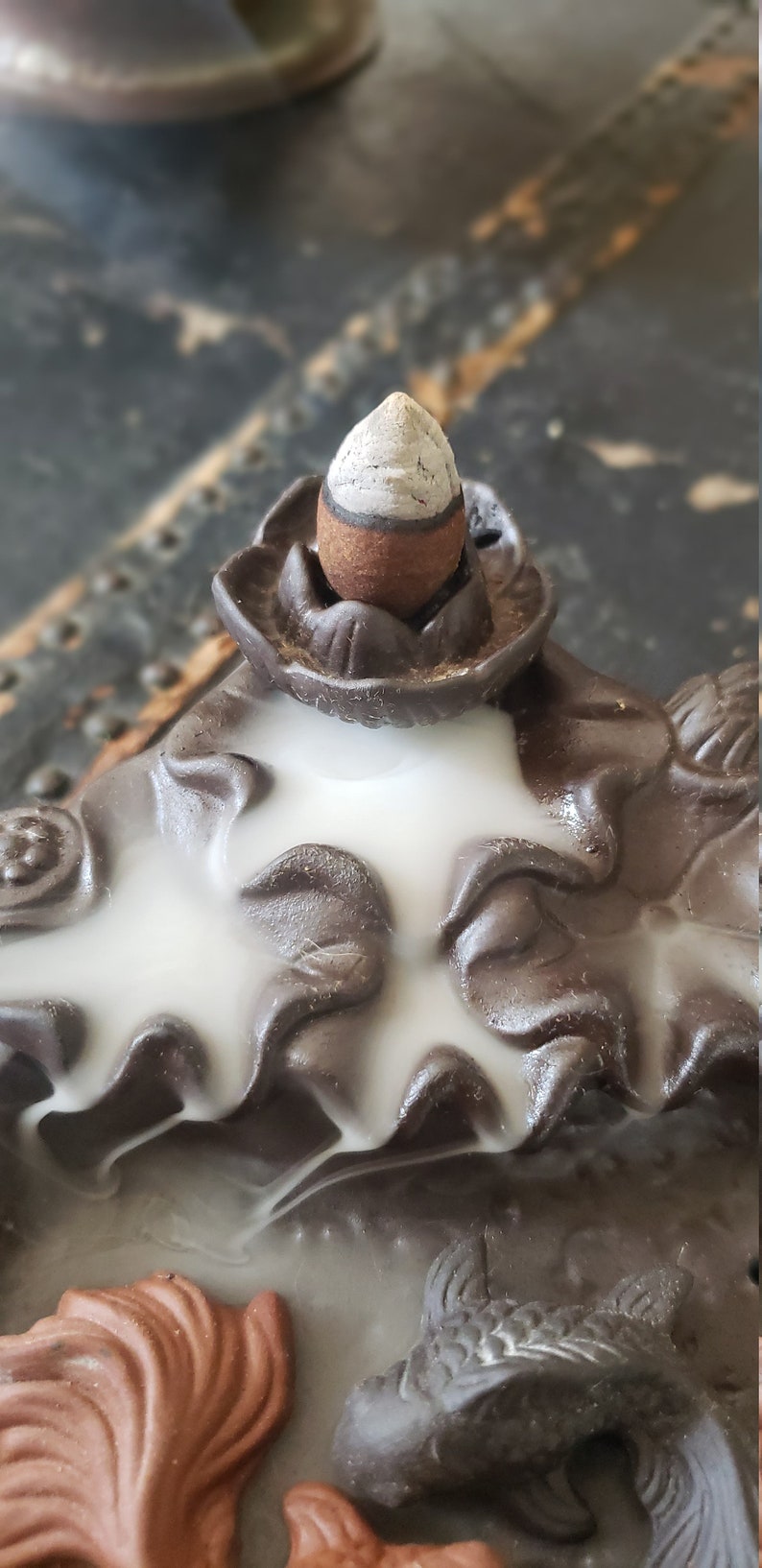 Peut inclure: Un br&ucirc;leur d'encens en c&eacute;ramique brune avec un motif de fum&eacute;e blanche. Le br&ucirc;leur pr&eacute;sente un motif de fleur de lotus avec un porte-encens conique sur le dessus. Le br&ucirc;leur a &eacute;galement deux motifs de poissons sur le fond.
